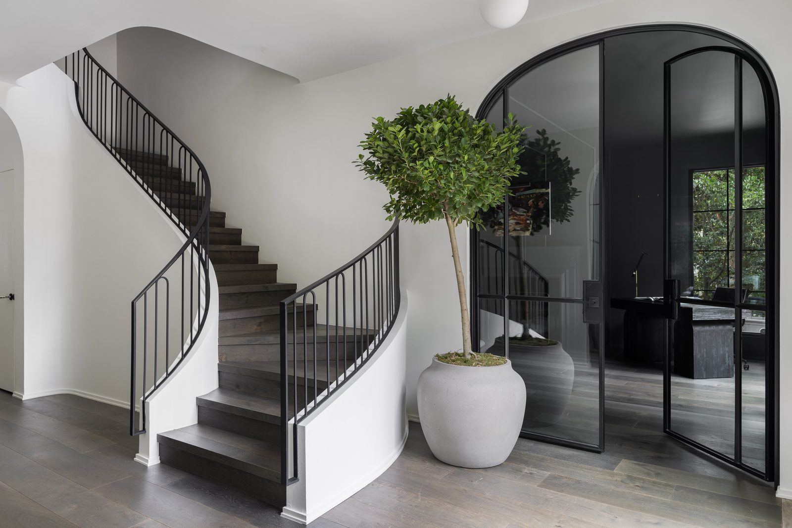 Editorial interior photograph of a curved staircase entry with steel-framed glass doors and a potted tree — the kind of final image agreed on shoot day, before the edit begins.