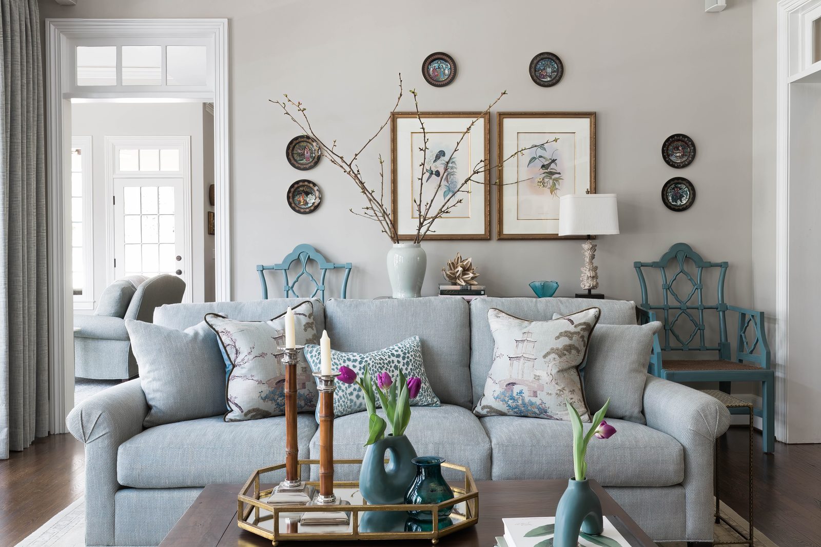 Wide editorial interior photograph of a soft gray living room with patterned blue armchairs, a watercolor diptych above the sofa, fresh tulips on a brass tray, a frame built around asymmetric composition and considered focal layering.