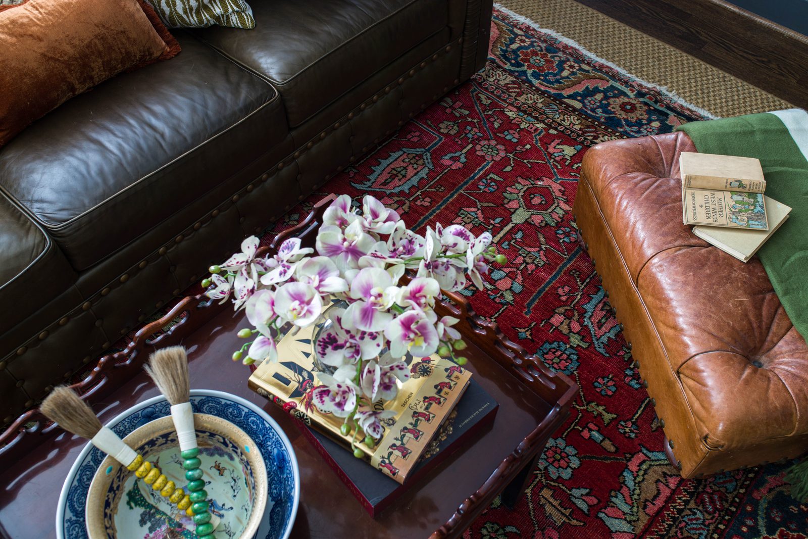 Overhead detail vignette of orchids, vintage books, and a blue-and-white bowl styled on a coffee table beside a leather ottoman, too narrow for a project page, ideal as an Instagram grid tile.
