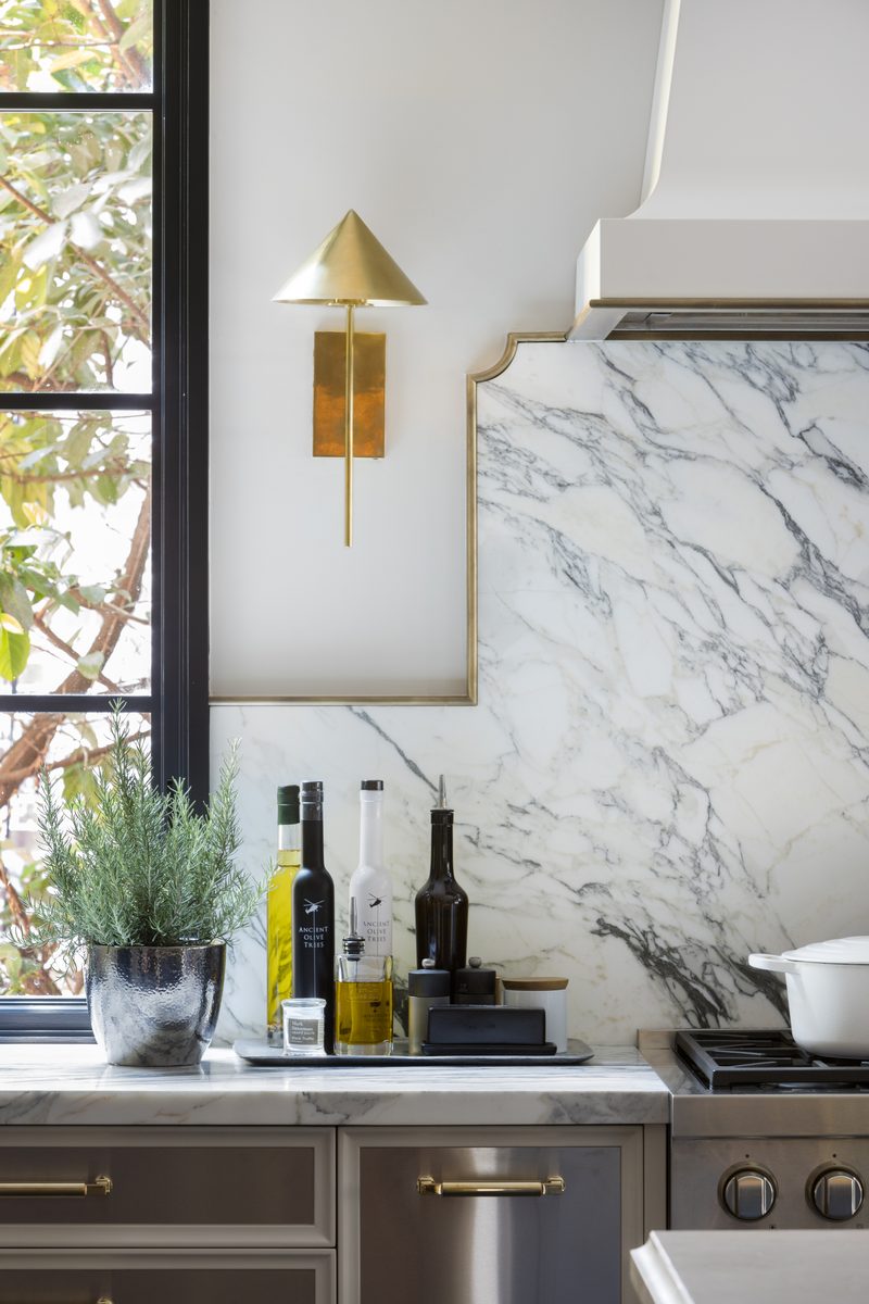 Tight kitchen vignette of a dramatic marble backsplash, brass sconce, and styled countertop by a steel window, a single-focal-point frame that stops a scroll.