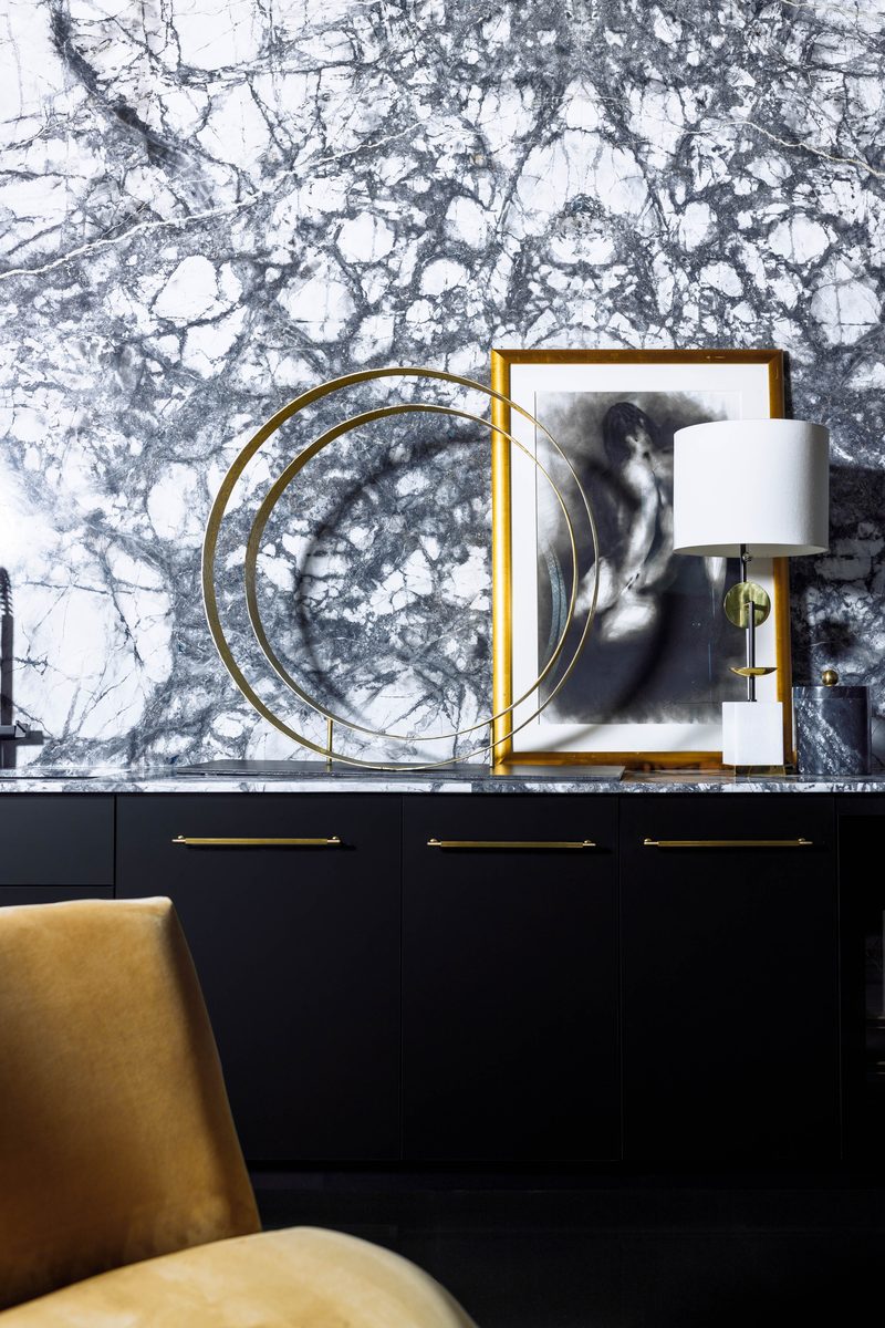 Vertical editorial detail of a console vignette: black marble waterfall counter, brass-ringed sculptural object, a framed black-and-white figure study, and a brass-and-marble lamp, demonstrating focal-point restraint and asymmetric framing.
