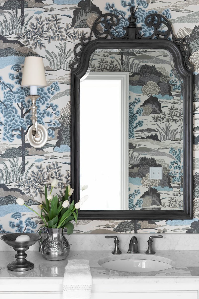 Vertical editorial detail of a powder room: a dark-framed ornate mirror reflecting a window, an antique-style sconce, white tulips in a pewter pitcher on a marble counter, against a blue-and-cream scenic wallpaper, exactly the vertical-format frame shelter magazines use for lead spreads.