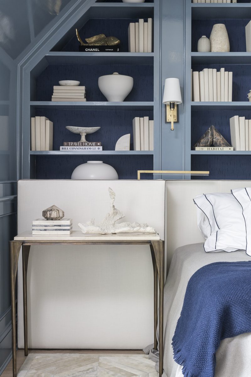 Vertical frame of a blue bedroom with lacquered built-in bookcases, a brass sconce, and a styled nightstand — the kind of vertical a features editor needs for cover-adjacent slots.