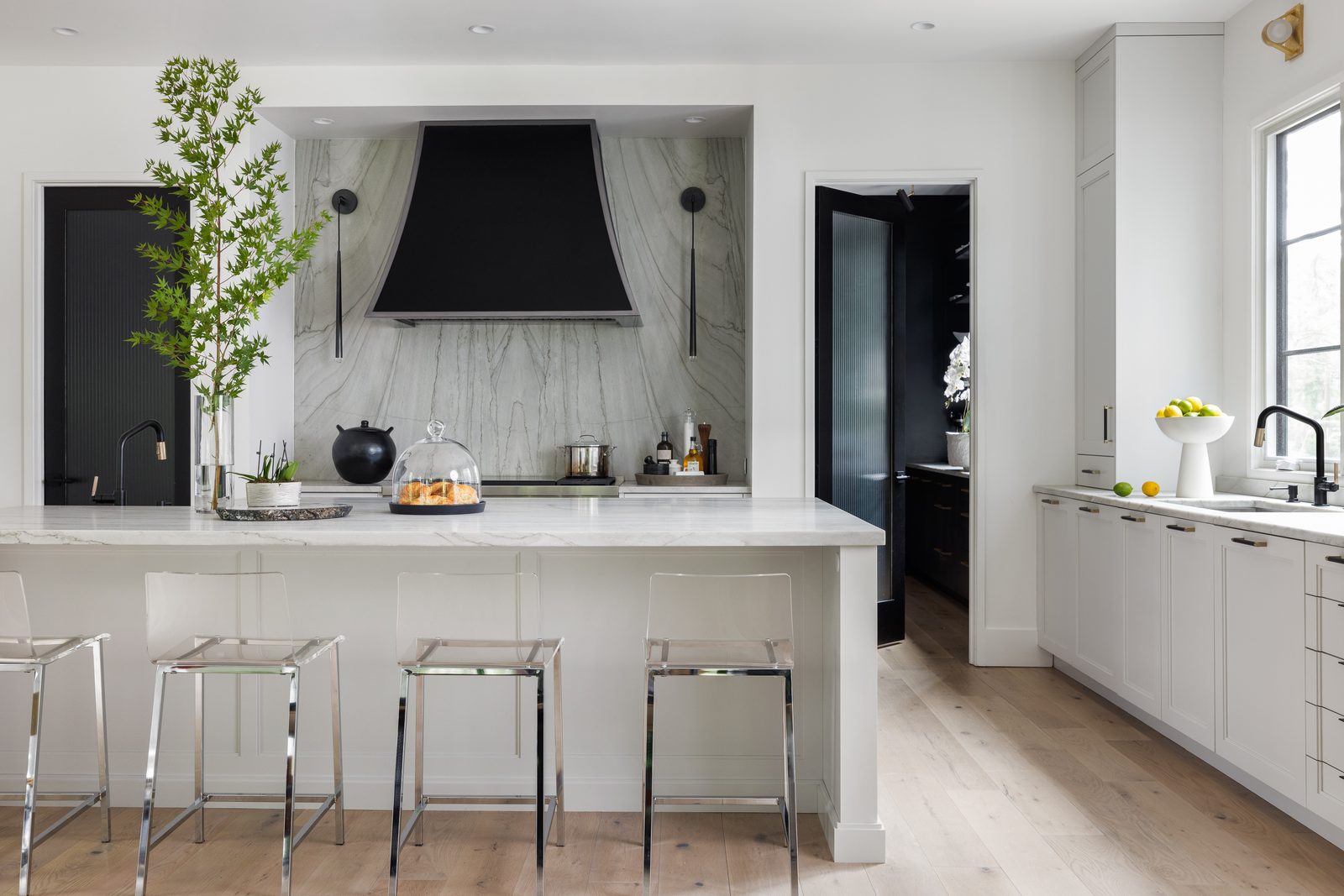 White kitchen with a black range hood, dramatic stone backsplash, and acrylic counter stools — composition built around the design story rather than a real estate listing.