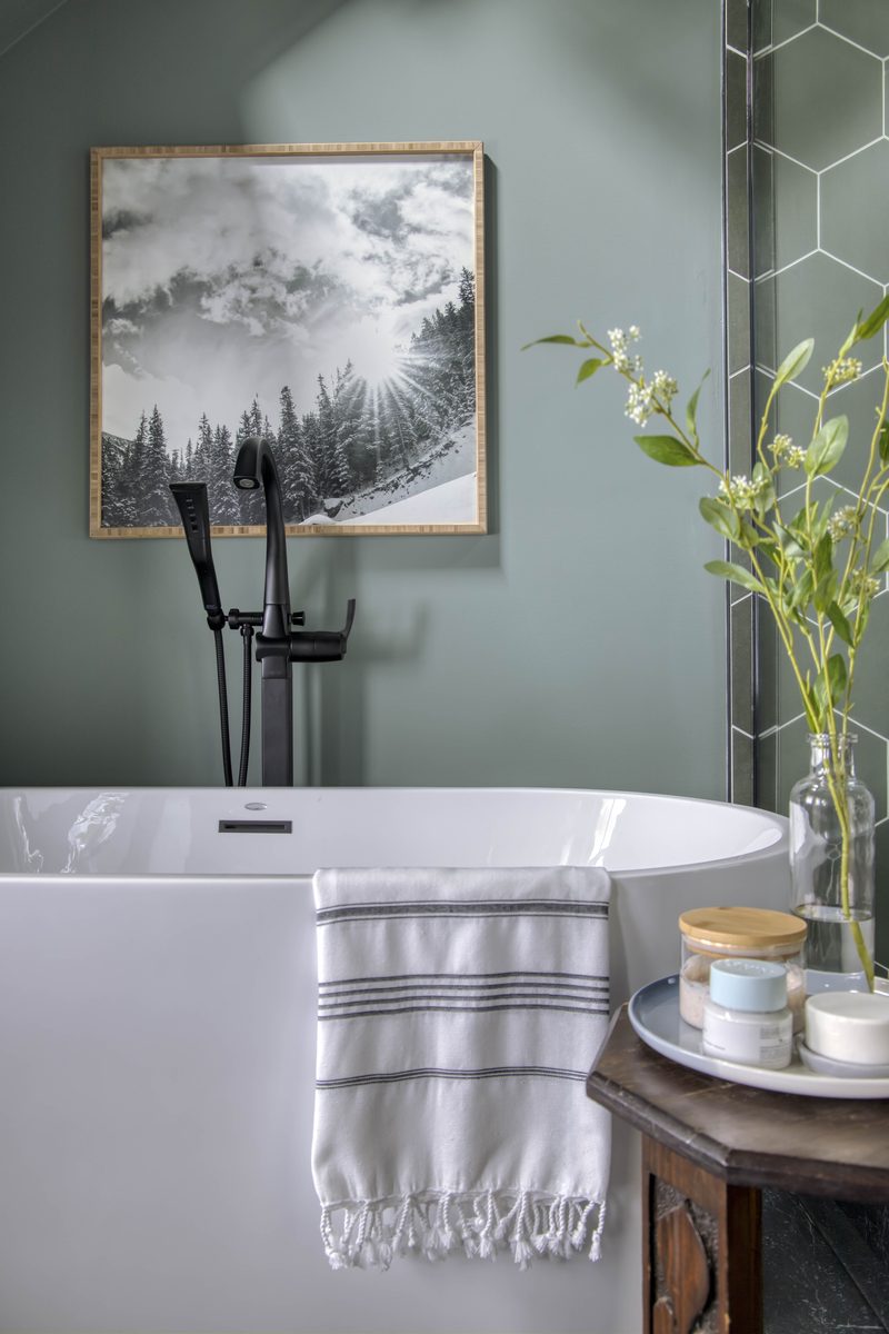 Vertical editorial detail of a freestanding tub with a fringed striped hand towel draped over the rim, fresh florals in a glass vase, and a framed landscape print: the textile and surface styling decisions that show up immediately through the lens.