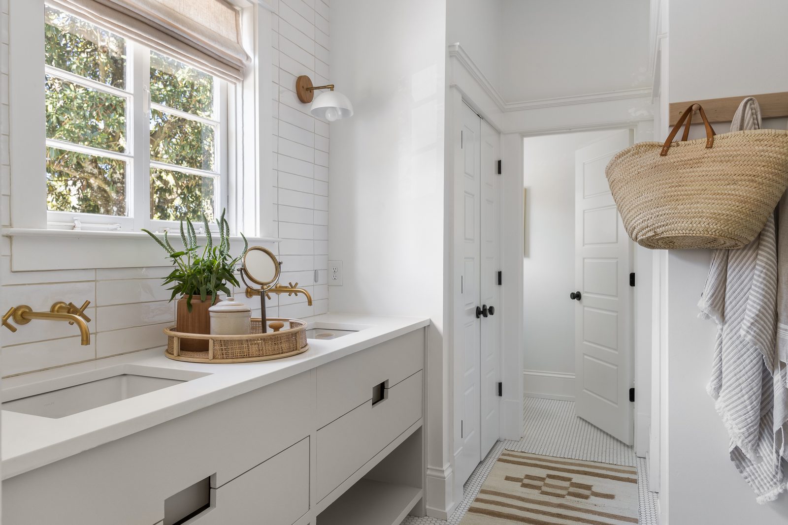 Styled white bathroom vanity with brass wall-mount faucets, fresh greenery, and a market basket on the door — the kind of pared-back styling that gets finished in the two weeks before a shoot.