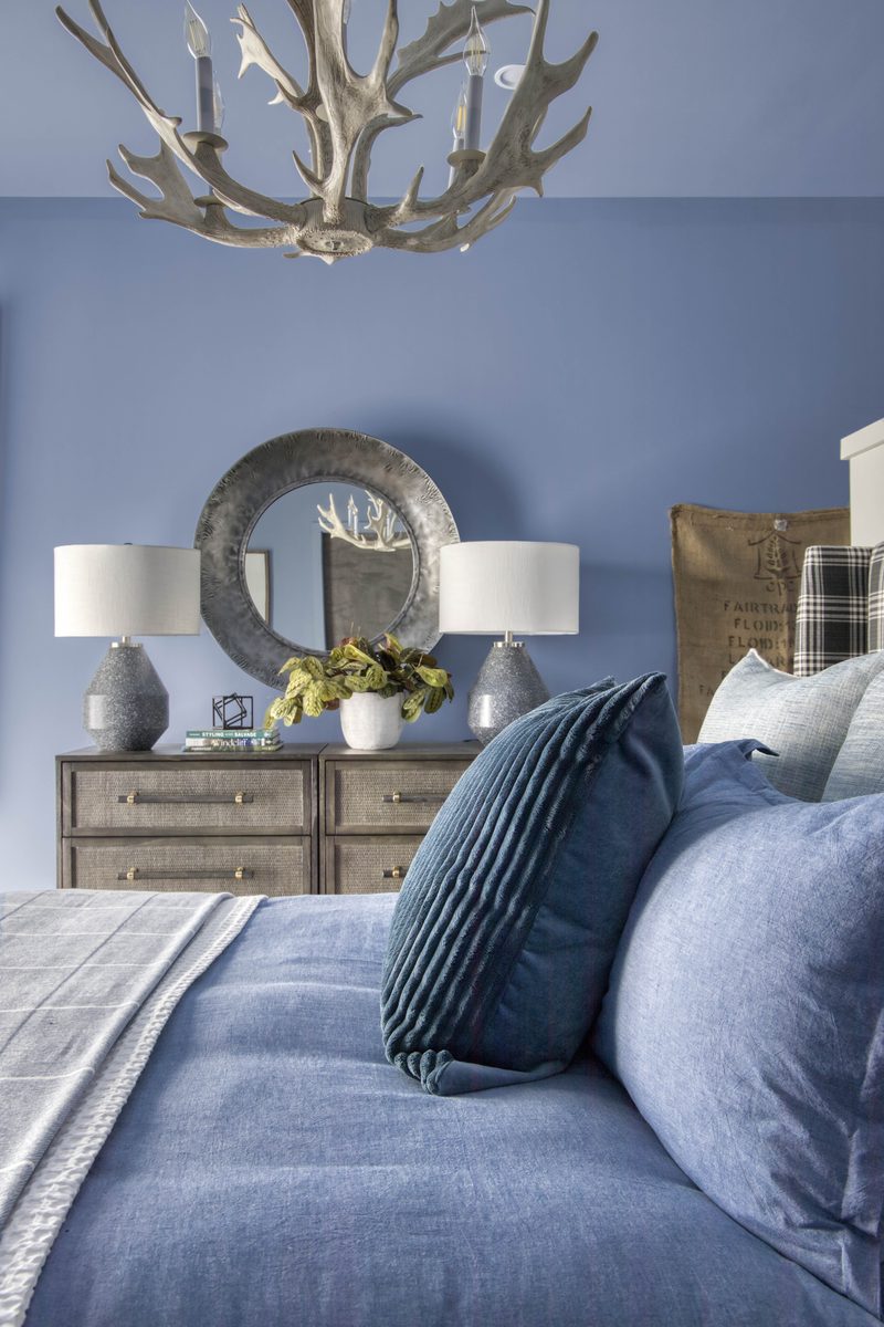 Vertical editorial interior photograph of a periwinkle bedroom with an antler chandelier, twin lamps on a textured dresser, a round mirror, and layered blue and tartan bedding: a textile-heavy frame where every styling decision is visible to the camera.