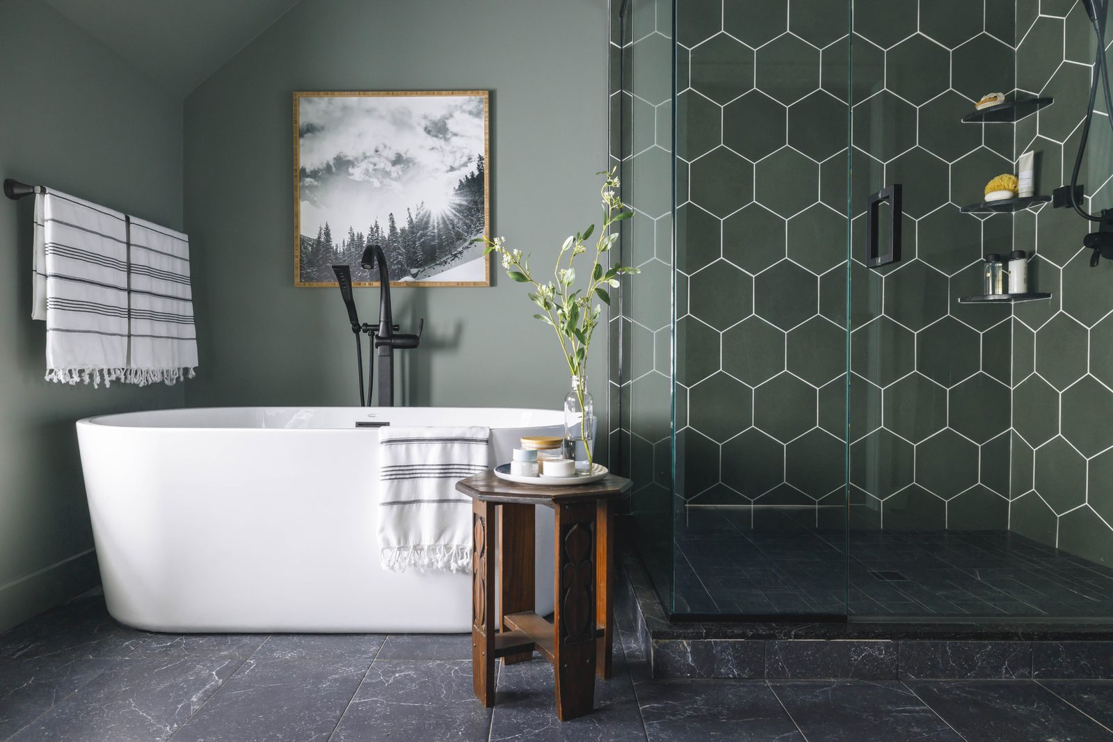 Editorial interior photograph of a primary bathroom with sage-green hexagonal tile, a freestanding tub styled with a striped hand towel, framed landscape art, and florals on a small side table: every visible element is a styling decision.