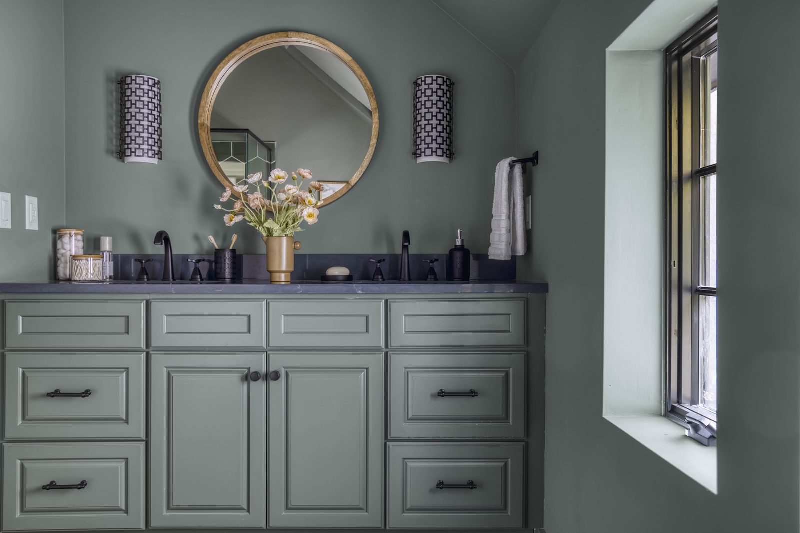 Editorial interior photograph of a sage-green powder room vanity with a round mirror, twin patterned sconces, a single arrangement of cream daisies, and dark stone counter: restrained styling with one focal point, the visual equivalent of 'when in doubt, take it away'.