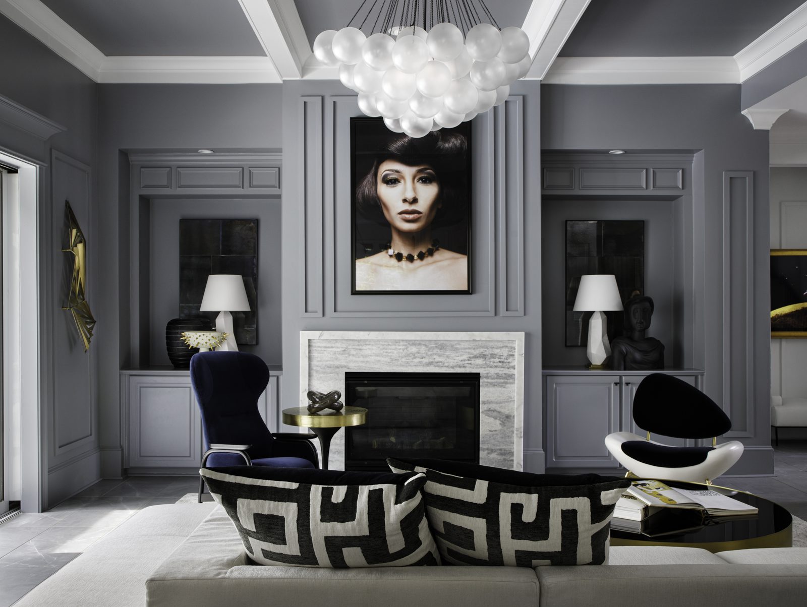 Editorial interior photograph of a formal gray living room with a large portrait above a marble fireplace, geometric pillows, brass accent table, and a cluster pendant light, the kind of considered editorial composition that earns publication.