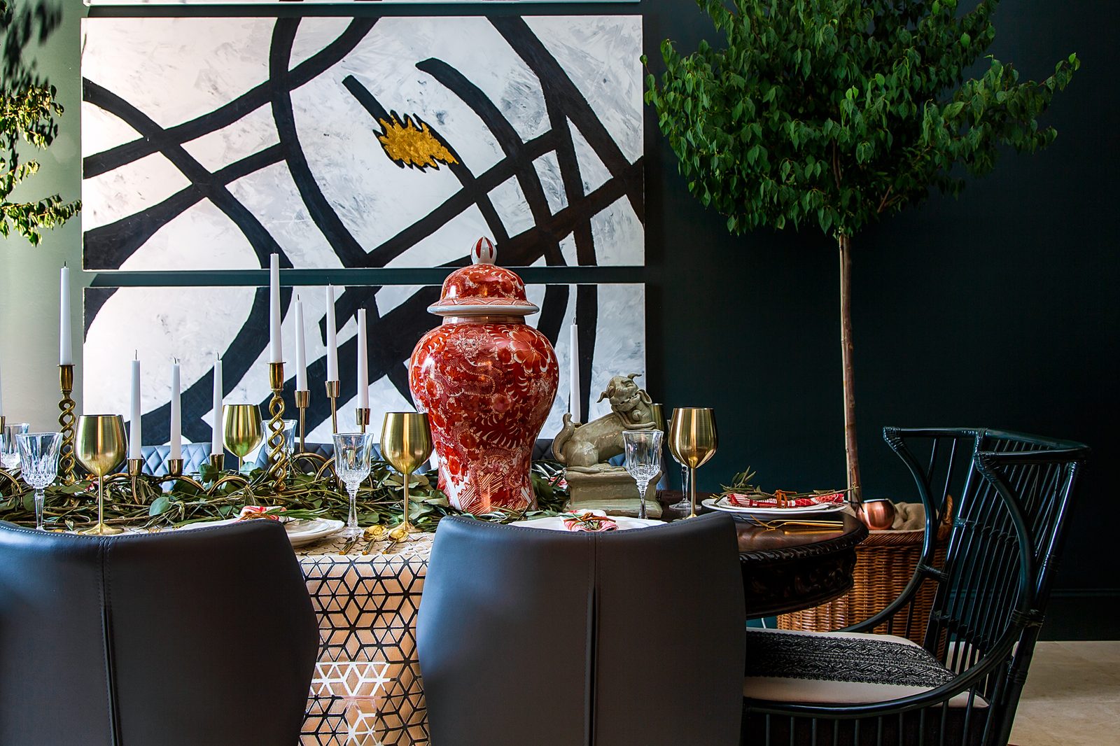 Moody dining room with a bold black-and-white mural, sculptural tree, and red ginger jar — a room composed around one clear story, the kind of frame that survives an editor's first pass.