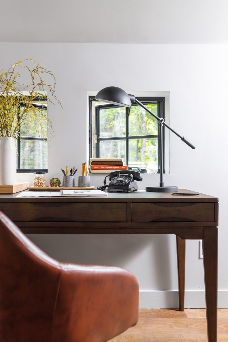 Vertical editorial photograph of a styled home office: a walnut desk with a vintage black telephone, brass desk lamp, books propped against a window, fresh greenery in a white vase, and a leather chair pulled in, a frame that reads as prepared down to the smallest surface detail.