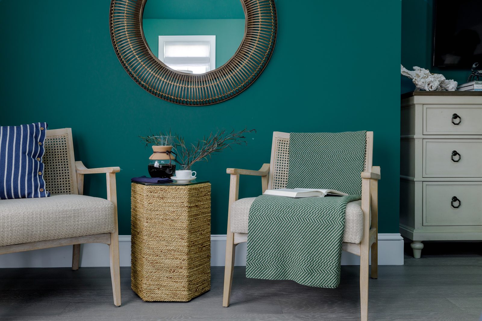Editorial interior photograph of a deep-teal sitting room with a round mirror, two woven-back chairs styled with a striped pillow and a green-and-cream throw, and a circular rattan side table holding a coffee press and bud vase, the kind of composed, momentum-built frame a prepared shoot day produces.