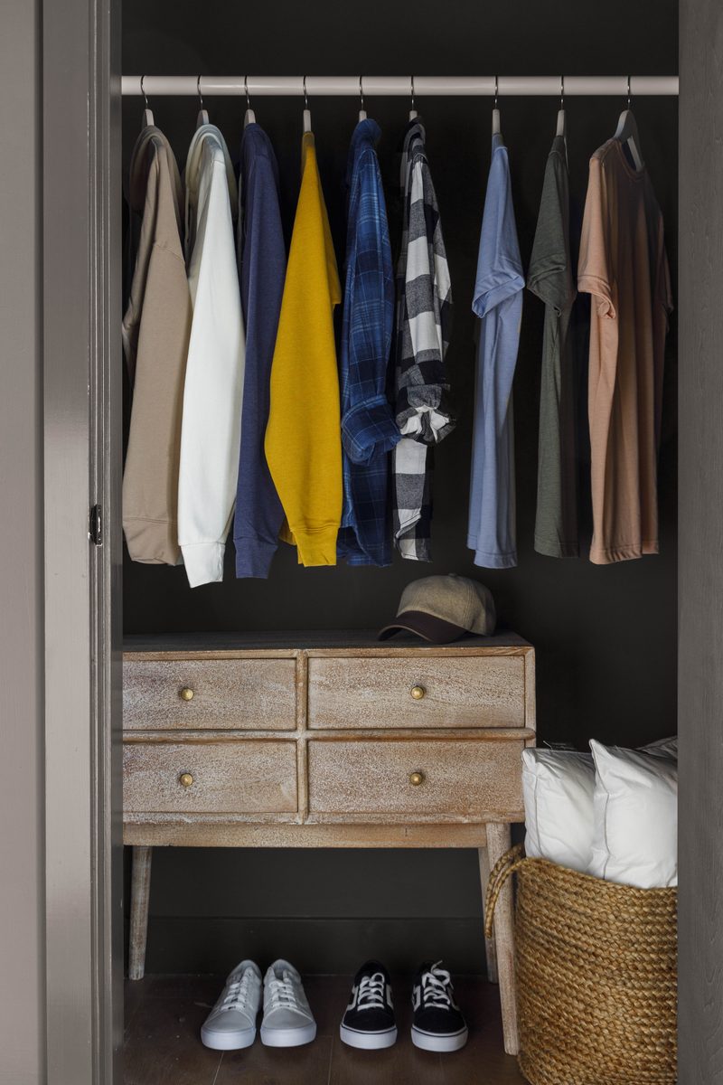 Vertical editorial photograph of an open custom closet: a row of intentionally arranged garments on matching hangers, a four-drawer console with brass pulls, a folded blanket and woven basket below, and two pairs of sneakers on the floor, visual shorthand for the kind of organized infrastructure a well-built shoot kit represents.