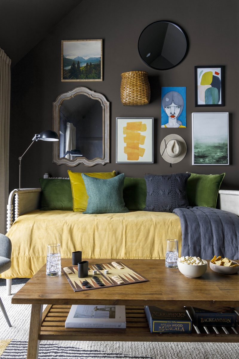 Vertical editorial photograph of a sitting room with charcoal walls, a gallery of mixed art and a woven basket above a yellow-upholstered daybed, layered with green, mustard, and slate pillows and a charcoal throw: exactly the kind of textile-heavy frame where steamed fabric makes or breaks the image.