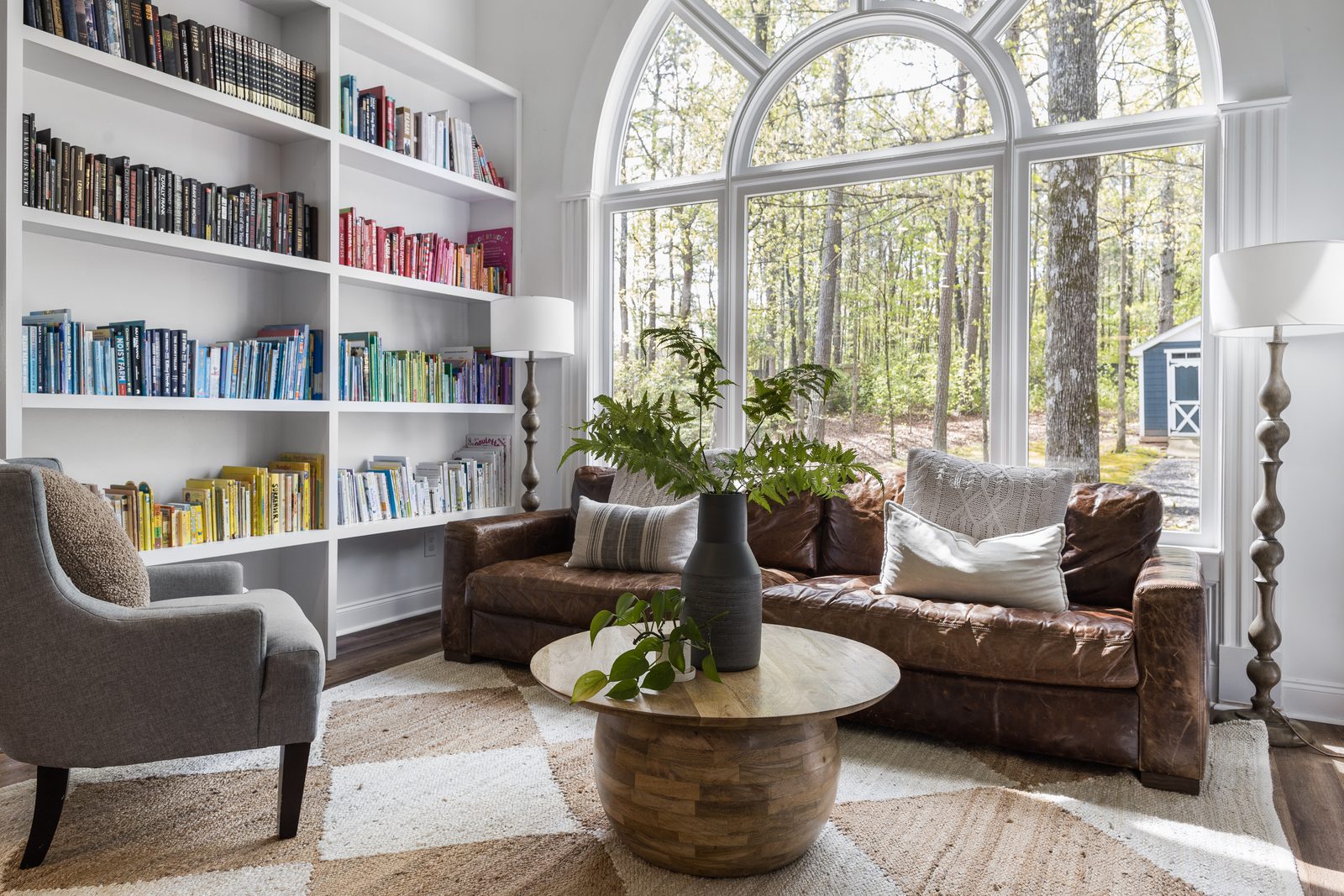 Sunlit reading room with a dramatic arched window, color-arranged built-in bookshelves, and a leather sofa — the kind of finished, styled space a planned shoot is built around.